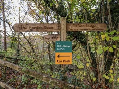 A wooden signpost with directions to Mardon Down Fingle Bridge Clifford Bridge and a sign for Clifford Bridge Car Park in a wooded area
