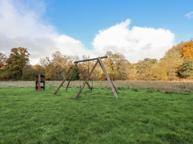 A playground with a wooden swing set and climbing structure on grass surrounded by trees at Dartmoor Retreat Lodge in Clifford Bridge near Moretonhampstead