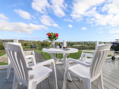 A patio with white plastic chairs a round table with flowers and cups and a barbecue grill on a deck with a view of green fields at Chestnut Lodge in Teigngrace near Newton Abbot