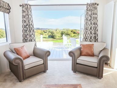 Two armchairs with cushions facing a large open window with a view of outdoor chairs and greenery at Chestnut Lodge in Teigngrace near Newton Abbot