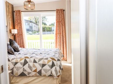 A bedroom with a bed near a large window with orange curtains and a view of green grass and houses at Chestnut Lodge in Teigngrace near Newton Abbot