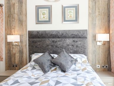A bed with a patterned duvet and two pillows with a textured headboard two framed pictures and wall lamps in a bedroom at Chestnut Lodge in Teigngrace near Newton Abbot
