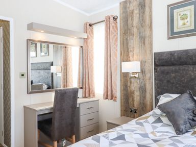A bedroom with a bed a bedside table a chair a desk with drawers a large mirror and patterned curtains at Chestnut Lodge in Teigngrace near Newton Abbot