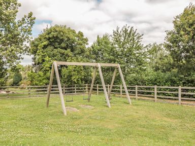 A wooden swing set with two regular swings and one baby swing on a grassy area surrounded by a wooden fence and trees at Chestnut Lodge Teigngrace near Newton Abbot
