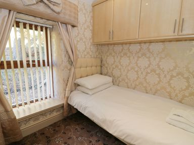 A small bedroom with a single bed pillows and towels beside a window with vertical blinds and wall cabinets at Bryn Berthen in Penrhyndeudraeth