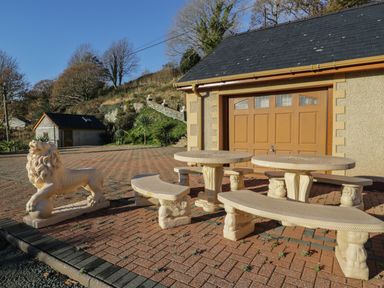 An outdoor area with stone lion statues and curved stone benches and tables in front of a building with a wooden garage door at Bryn Berthen in Penrhyndeudraeth