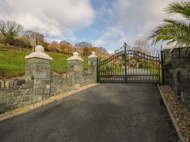 A paved driveway leading to a decorative metal gate with stone pillars and a green hillside in the background at Bryn Berthen in Penrhyndeudraeth