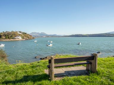 A bench on grass overlooking a body of water with boats and hills in the background at Bryn Berthen in Penrhyndeudraeth