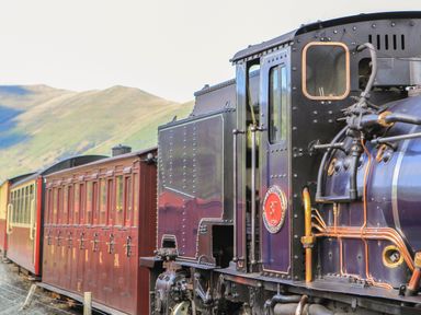 A steam locomotive pulling passenger train carriages with mountains in the background at Bryn Berthen in Penrhyndeudraeth