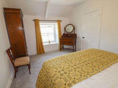 A bedroom with a bed covered by a patterned yellow blanket a wooden wardrobe a chair a wooden dressing table with a mirror curtains on a window and a closed door at Delfryn Newport in Newport Pembrokeshire