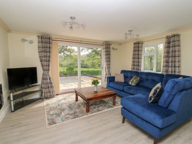 A living room with a blue sectional sofa a wooden coffee table a television on a stand and large windows with curtains at Erwlon in Rhydyfelin near Aberystwyth