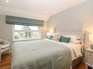 A bedroom with a bed green blanket green and patterned cushions a white bedside table lamp a window with a green valance and a wicker chair at Fron Olau in Nefyn