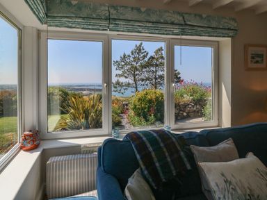 A living room with a blue sofa and cushions near large windows showing a garden and sea view at Fron Olau in Nefyn