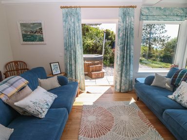 A sitting room with two blue sofas, patterned curtains, a rug, and a door opening to a garden with a hot tub at Fron Olau in Nefyn