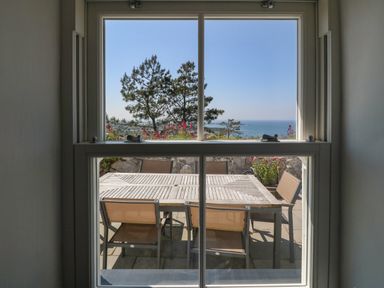 View through a window of an outdoor dining table and chairs with trees and sea in the background at Fron Olau in Nefyn