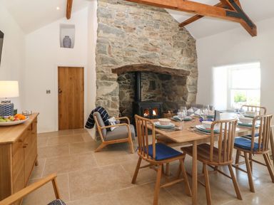 A dining room with a stone fireplace and wood burning stove a wooden dining table with chairs and blue cushions a wooden sideboard with a lamp and fruit bowl at Fron Olau in Nefyn