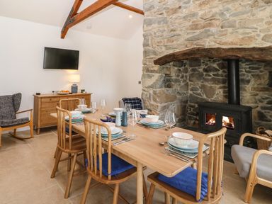 A dining room with a wooden table set for six near a stone fireplace with a wood stove and a wall-mounted TV at Fron Olau in Nefyn