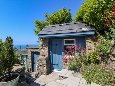 A stone building with blue doors surrounded by plants and flowers with an ocean view at Fron Olau in Nefyn