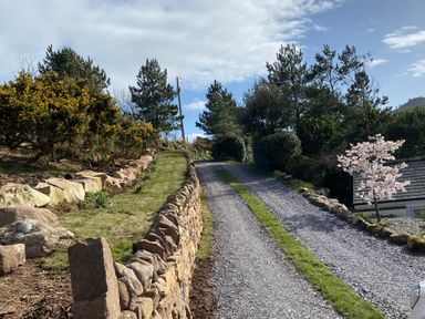 A gravel driveway with grass strips bordered by stone walls and trees at Fron Olau in Nefyn