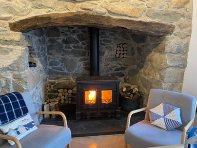A stone fireplace with a wood burning stove firewood stored inside and two armchairs with cushions in a living room at Fron Olau in Nefyn