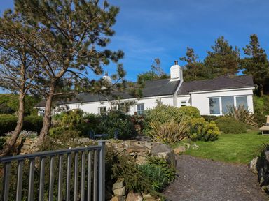 A white house with a slate roof surrounded by bushes and trees with a stone path and metal railing in the foreground at Fron Olau in Nefyn