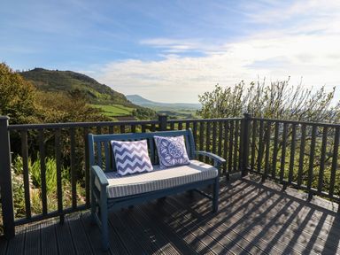 A blue bench with two cushions on a wooden deck overlooking a landscape with hills and trees at Fron Olau in Nefyn