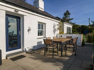 An outdoor patio area with a wooden table and six chairs next to a white building with a blue door and windows at Fron Olau in Nefyn