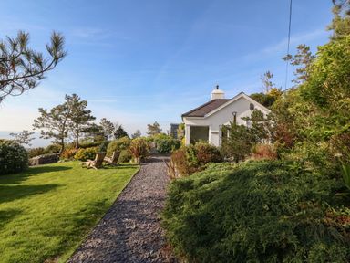 A white house with a slate roof next to a gravel path and green garden with chairs at Fron Olau in Nefyn