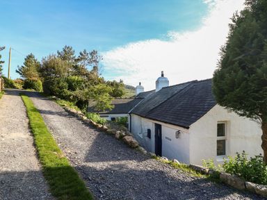 A gravel driveway with grass strip leading to a white house with dark roof and chimneys by trees and bushes at Fron Olau in Nefyn