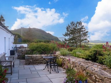 A stone patio with outdoor furniture and plants surrounded by stone walls and trees and hills in the background at Fron Olau in Nefyn