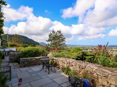 A stone patio with metal and wooden chairs and a table overlooking a garden with trees and flowers at Fron Olau in Nefyn