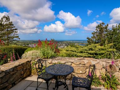 An outdoor patio with a metal table and two chairs overlooking a garden and distant coastline at Fron Olau in Nefyn