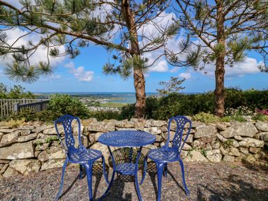 A blue metal table and two matching chairs on a gravel patio with trees and a stone wall overlooking a town and sea at Fron Olau in Nefyn