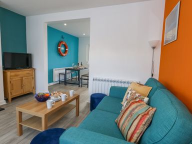 A living room with a blue sofa and colorful pillows a wooden coffee table with cups and fruit a TV on a wooden stand and a dining area with a life ring on the wall at Lagoon View in Wyke