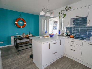 A kitchen with white cabinets, blue tiled backsplash, an island with a bowl of fruit and pastries, and a dining table with a bench against a blue wall with a life ring at Lagoon View in Wyke