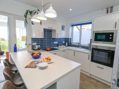 A kitchen with white cabinets blue tiled backsplash a white countertop with stools and appliances at Lagoon View in Wyke