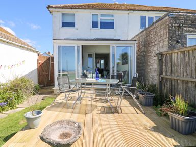 A backyard deck with a glass table and four chairs in front of a two-story house with sliding glass doors at Lagoon View in Wyke