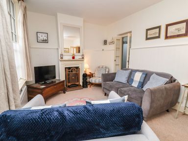 A living room with a grey sofa blue cushions a TV on a wooden chest a fireplace and an armchair at Tuckaway Cottage in Aldeburgh
