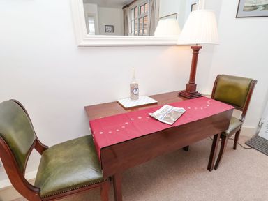 A wooden table with a red runner a newspaper a lamp and two green leather chairs beneath a wall mirror at Tuckaway Cottage in Aldeburgh
