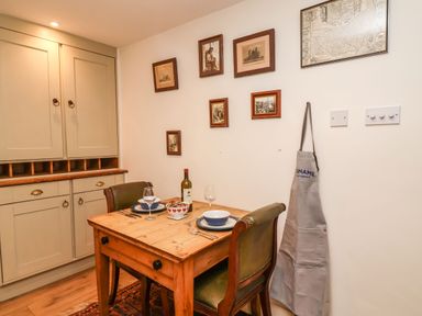 A dining area with a wooden table set for two, chairs, wall art, storage cabinets and an apron hanging on the wall at Tuckaway Cottage in Aldeburgh