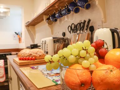 A kitchen counter with a glass bowl of fruit including grapes and oranges a loaf of bread and cherry tomatoes on a cutting board toasters knives hanging on a wall and blue mugs at Tuckaway Cottage in Aldeburgh