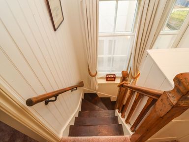 A narrow wooden staircase with dark steps and a wooden handrail beside a window with cream curtains at Tuckaway Cottage in Aldeburgh