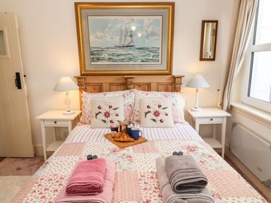 A bedroom with a bed with floral bedding and towels on it two bedside tables with lamps a window and a painting above the headboard at Tuckaway Cottage in Aldeburgh