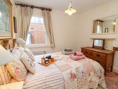 A bedroom with a bed covered in floral and striped bedding with pillows and towels on top and a wooden dresser with a mirror at Tuckaway Cottage in Aldeburgh