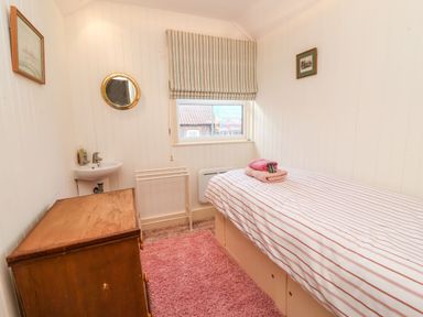 A small bedroom with a single bed a wooden chest of drawers a wall-mounted sink small mirror and a window with striped blinds at Tuckaway Cottage in Aldeburgh