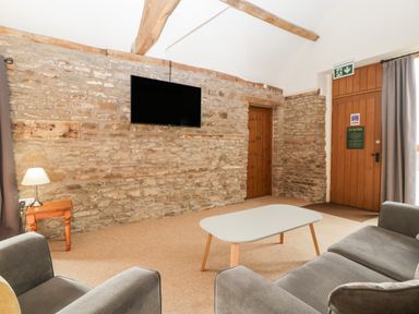 A living room with a stone wall a mounted television a wooden door a coffee table armchairs and a side table with a lamp at Peartree Cottage in Stanford Bishop near Bromyard