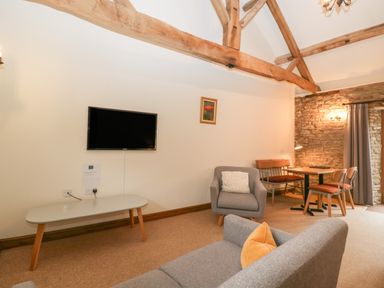 A living room with a wall-mounted television a gray armchair a gray sofa with a yellow cushion a dining table with chairs and a bench exposed wooden beams and a stone wall at Clover Patch Cottage in Stanford Bishop near Bromyard