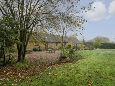 A garden with trees and picnic tables next to a stone building at Clover Patch Cottage in Stanford Bishop near Bromyard