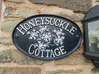 A black oval sign with white letters reading Honeysuckle Cottage mounted on a stone wall next to a lantern