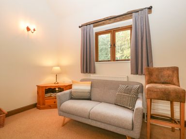 A small sitting room with a gray sofa, two cushions, a wooden corner table with a lamp, a window with gray curtains, and a brown bar stool at Honeysuckle Cottage Stanford Bishop near Bromyard